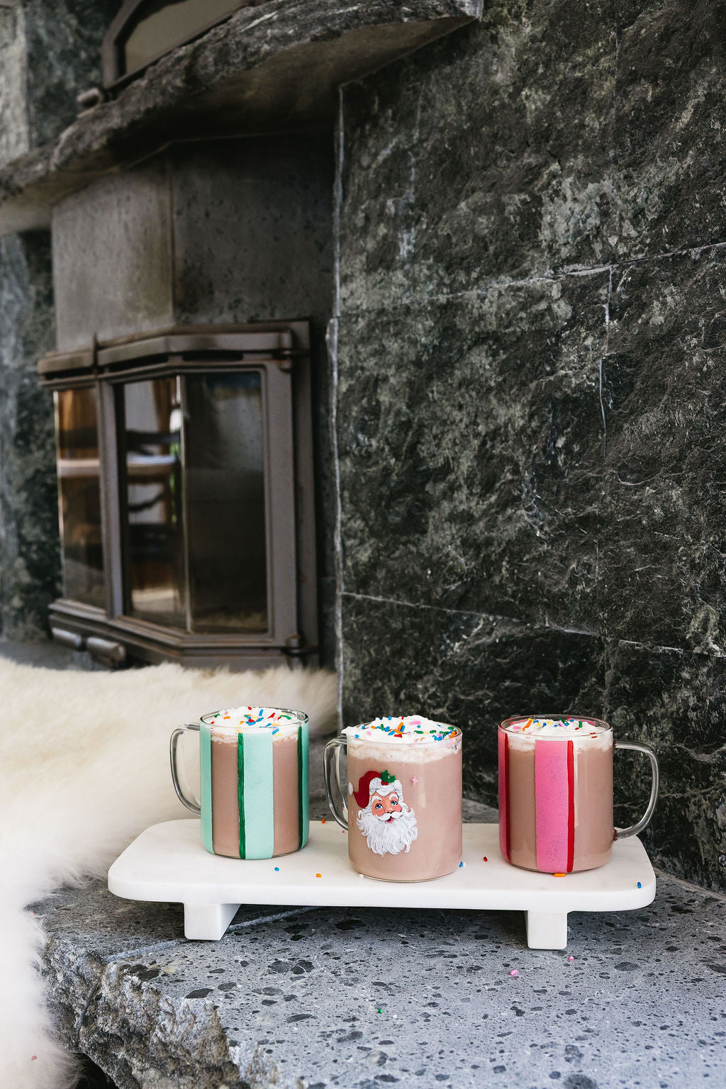 Three colorful mugs with festive designs on a white tray in front of a fireplace.